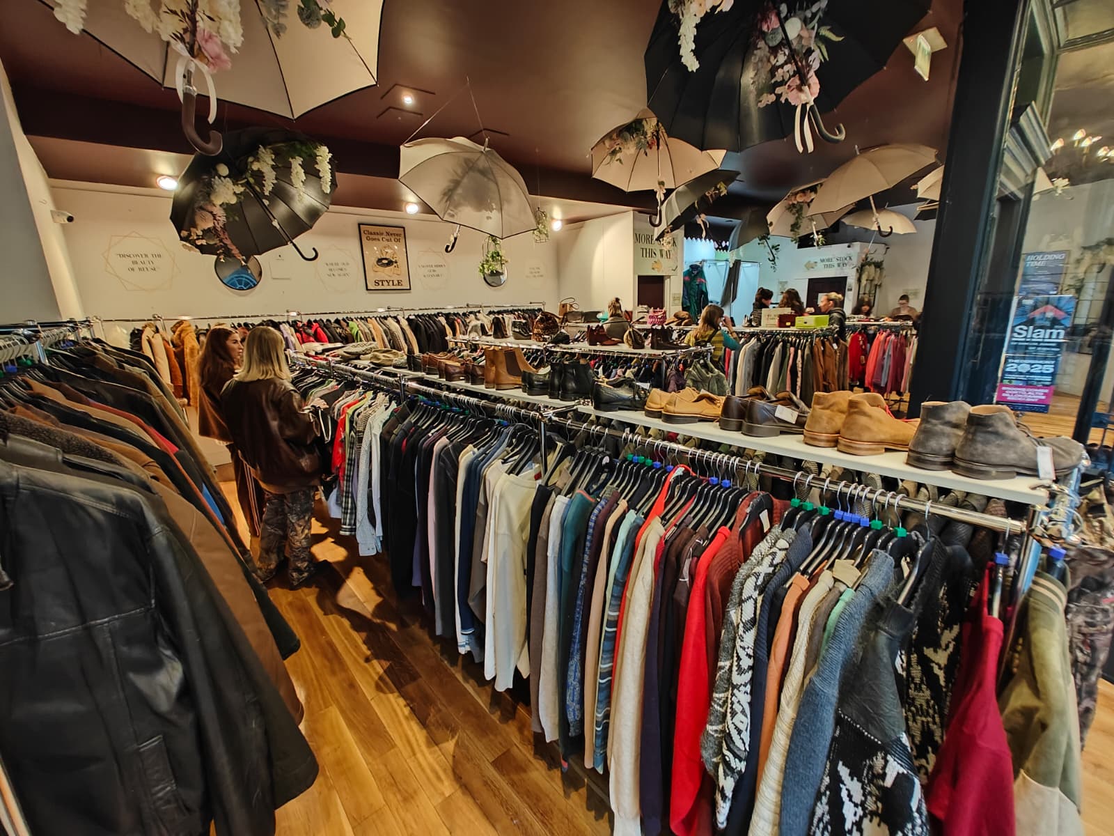 The inviting interior of Preloved Secret Garden, a second-hand women's clothing shop in Dundee, Scotland. Wide-angle of colourful clothes racks and decorative umbrellas hanging from the ceiling