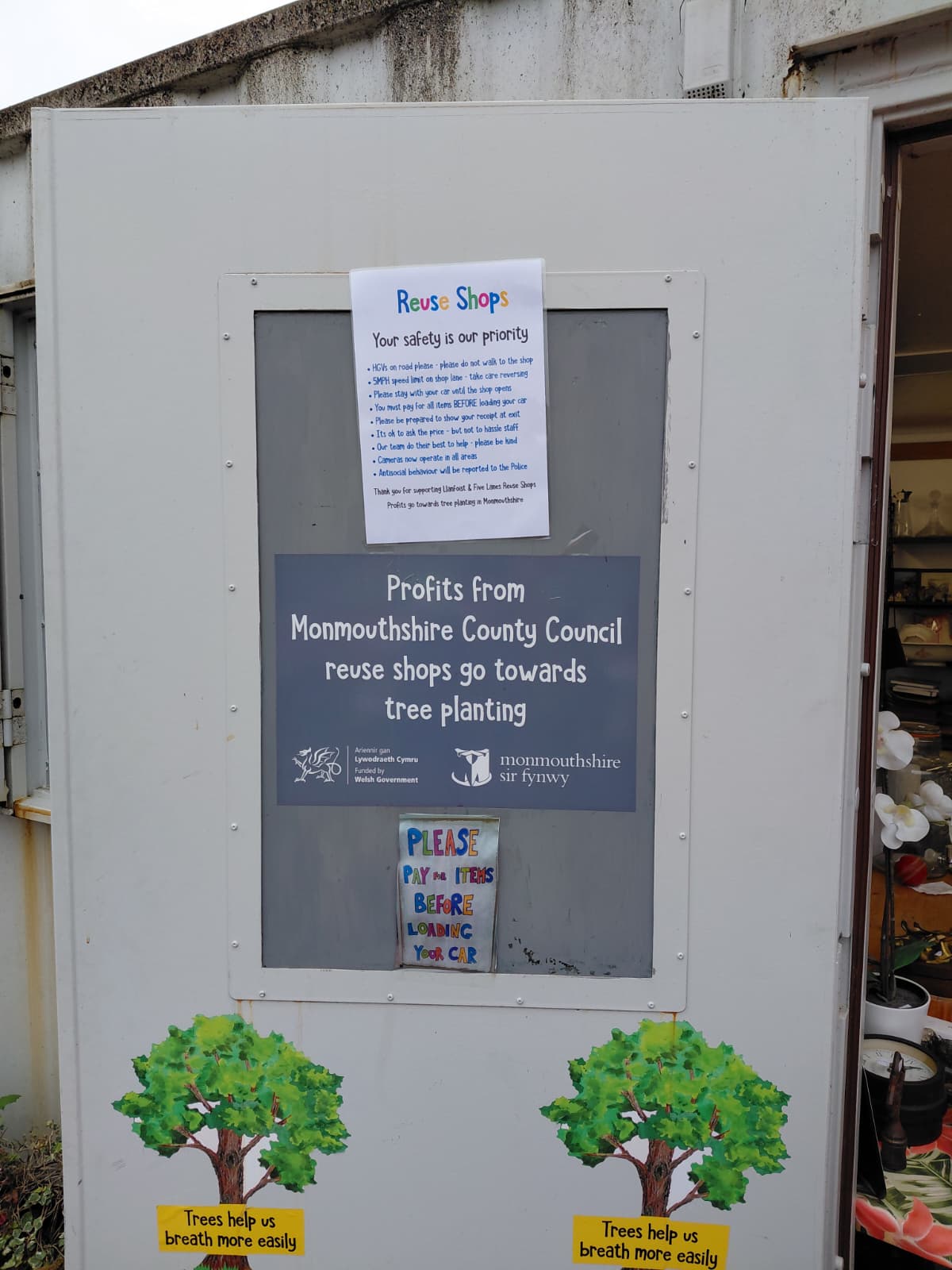 Entrance sign at a UK reuse shop: "Profits from Monmouthshire County Council reuse shops go towards tree planting" with safety notice and Welsh Government logos.