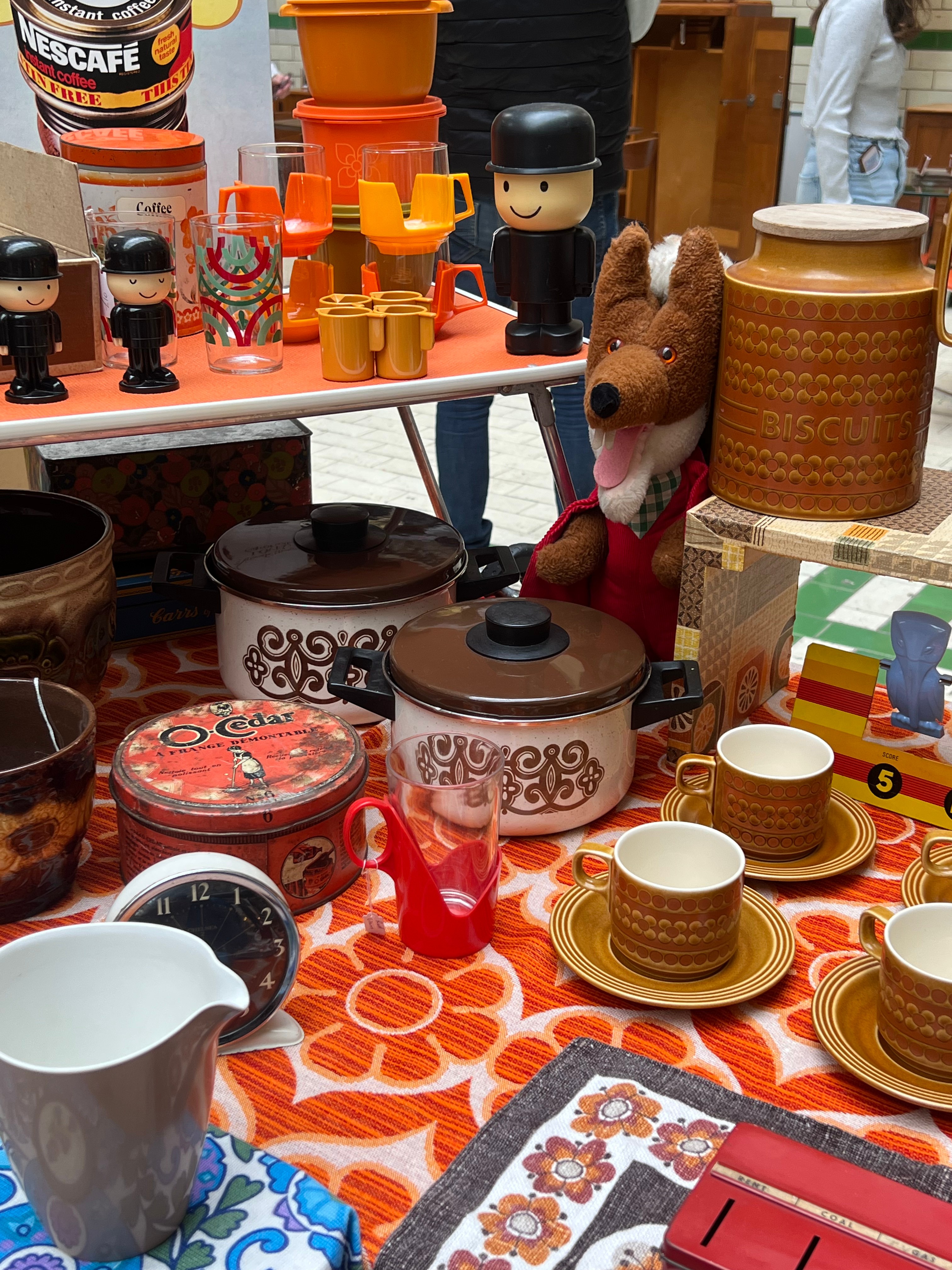 Close-up of a vintage market stall at the Vintage Home Show in Victoria Baths, Manchester, displaying retro kitchenware including 1970s crockery, patterned mugs, biscuit tins, enamel pots, and nostalgic toys on an orange geometric tablecloth.