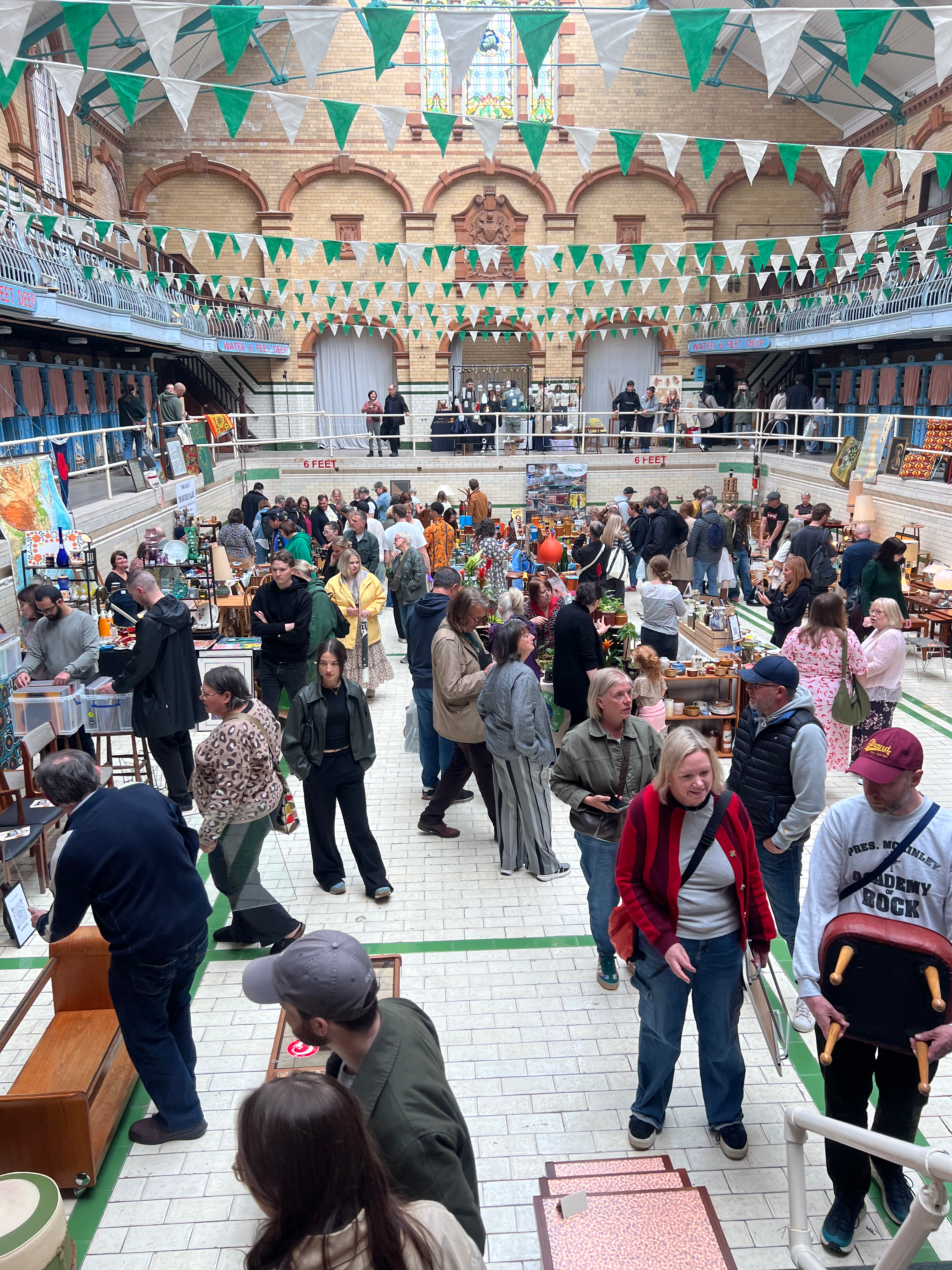 Shoppers exploring vintage stalls inside the drained Edwardian swimming pool at Victoria Baths during the Vintage Home Show in Manchester, surrounded by retro decor, and architectural heritage features like stained glass and tiled walls.