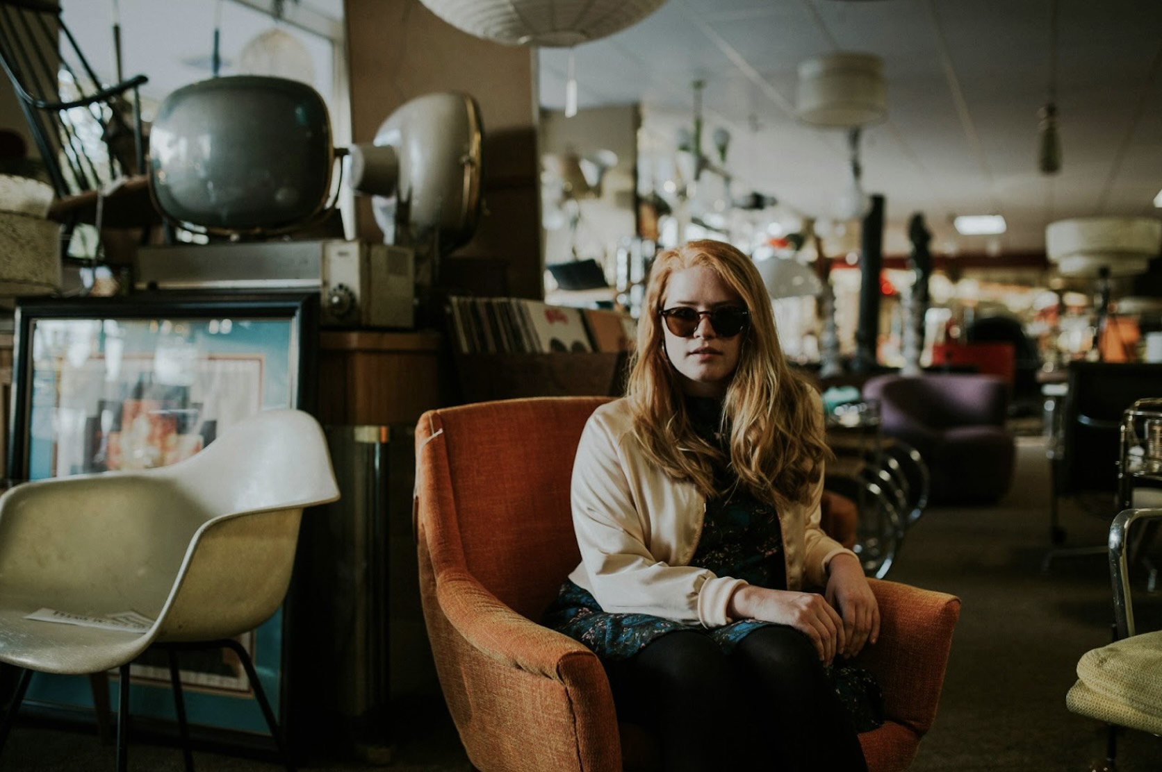 A woman looking very chic in sunglasses in a thrift store, sitting on an old chair