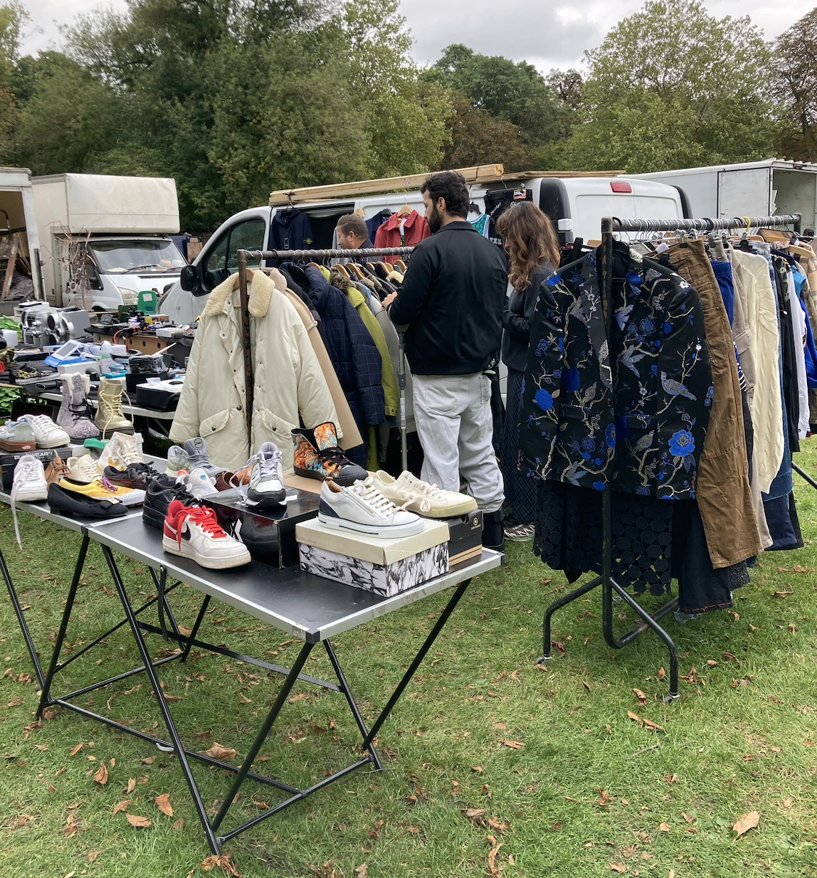 Stall at Chiswick Car Boot Sale displaying second-hand clothes, jackets, and trainers with sellers and shoppers browsing.
