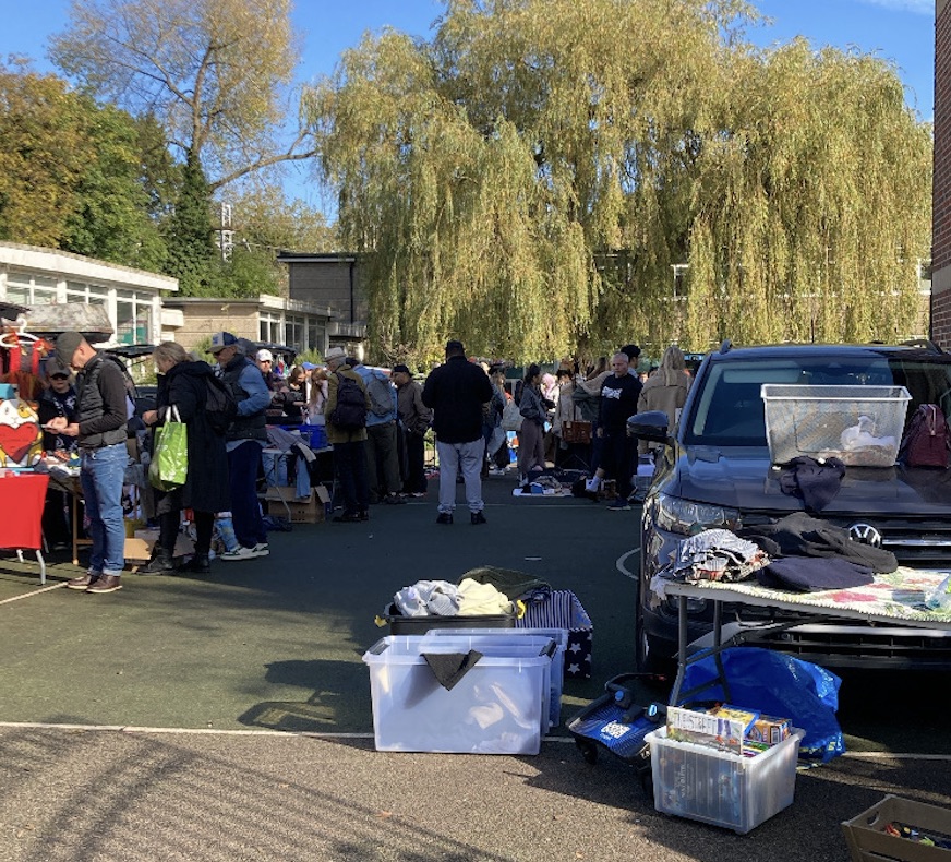 Outdoor Kentish Town car boot sale in London on a sunny day, with crowds browsing stalls, sellers displaying clothes and household items beside parked cars under large willow trees.