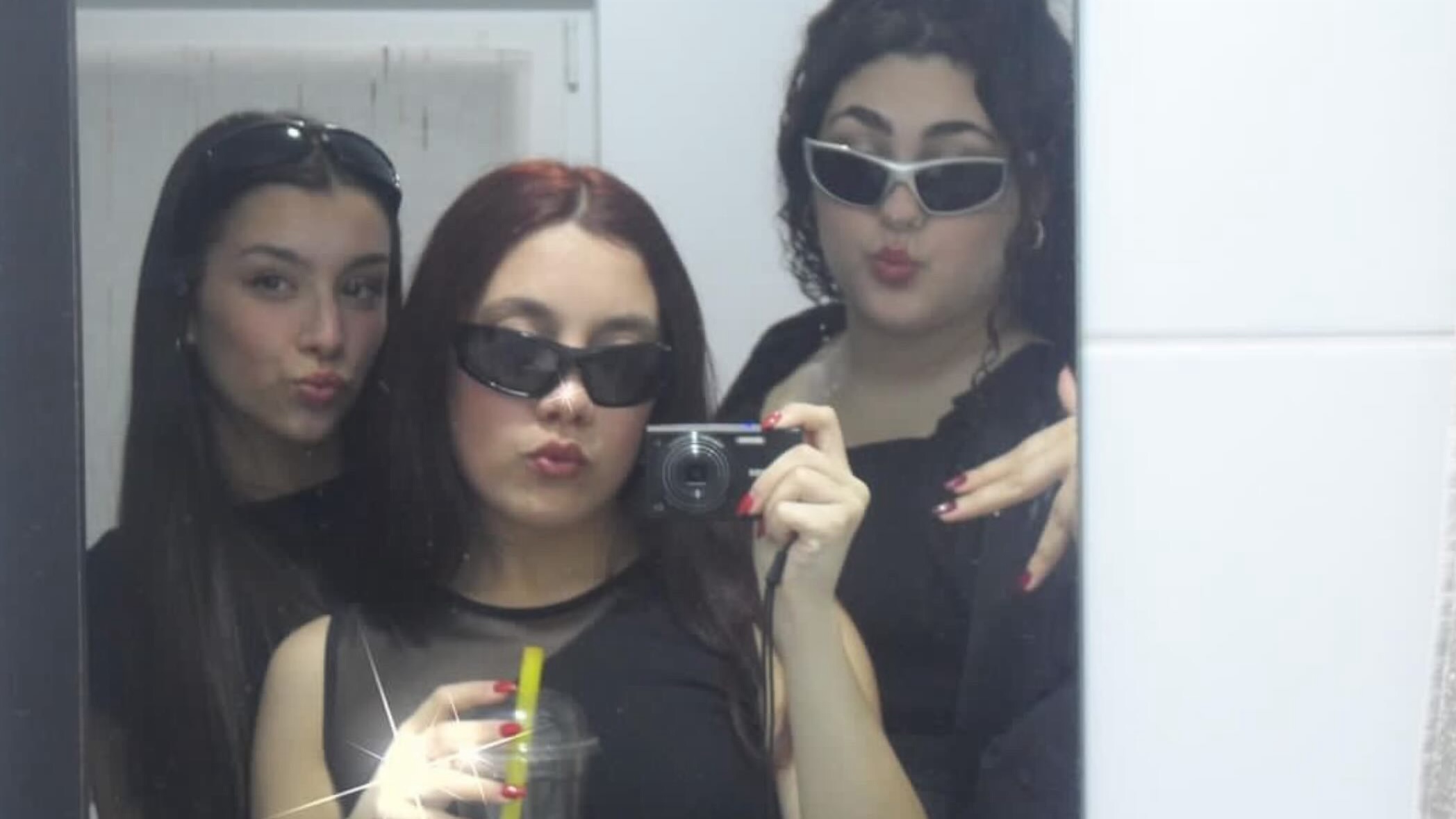 Three gen-z girls wearing y2k clothing and taking a group picture in a mirror using a vintage camera