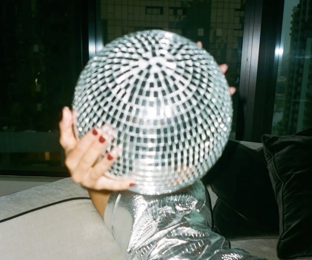 A woman wearing a vintage dress on a sofa and holding a disco ball at a party in London
