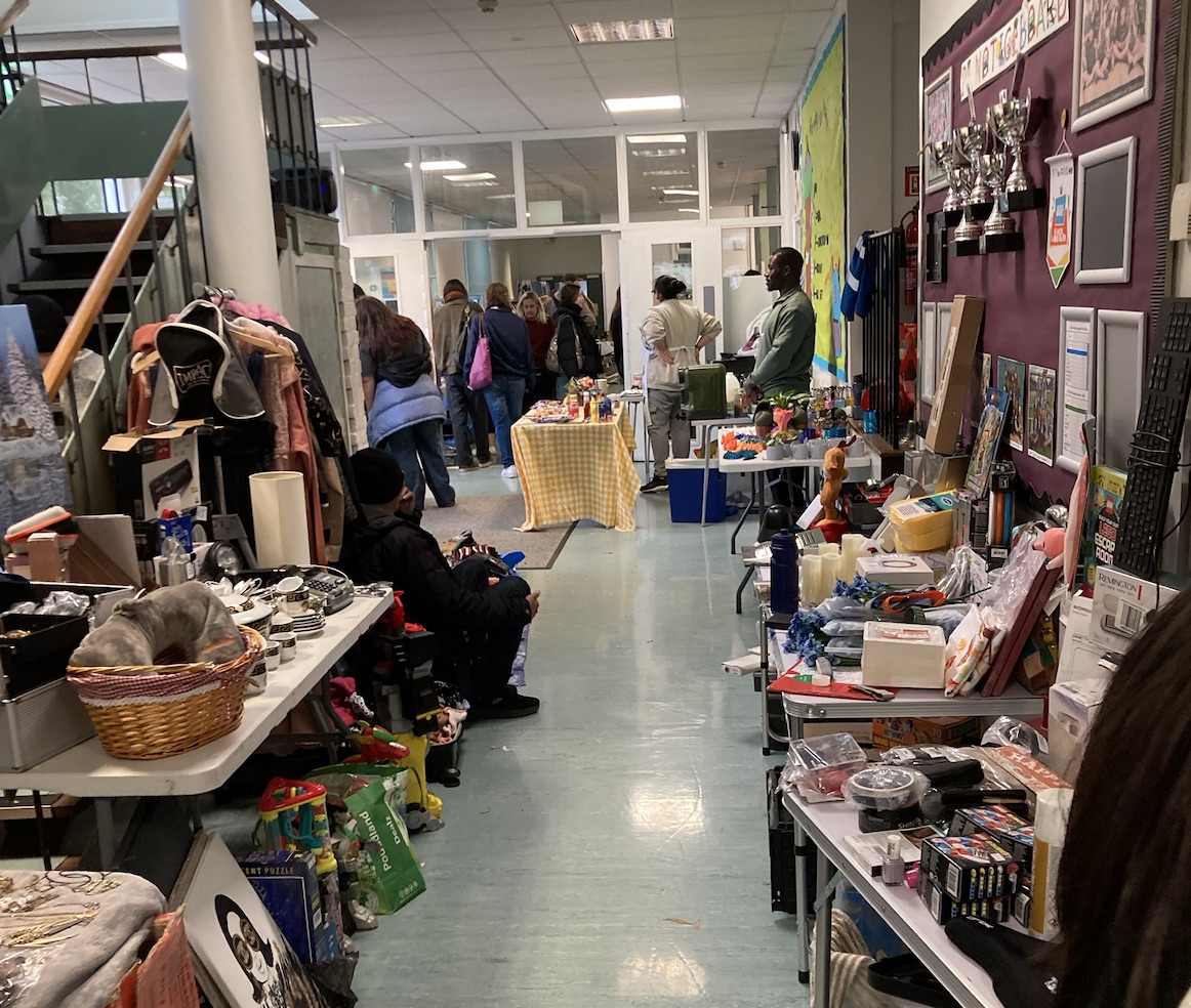 Indoor scene at Kentish Town car boot sale in London, showing tables filled with second-hand items, clothing racks, and shoppers browsing in a busy school corridor.
