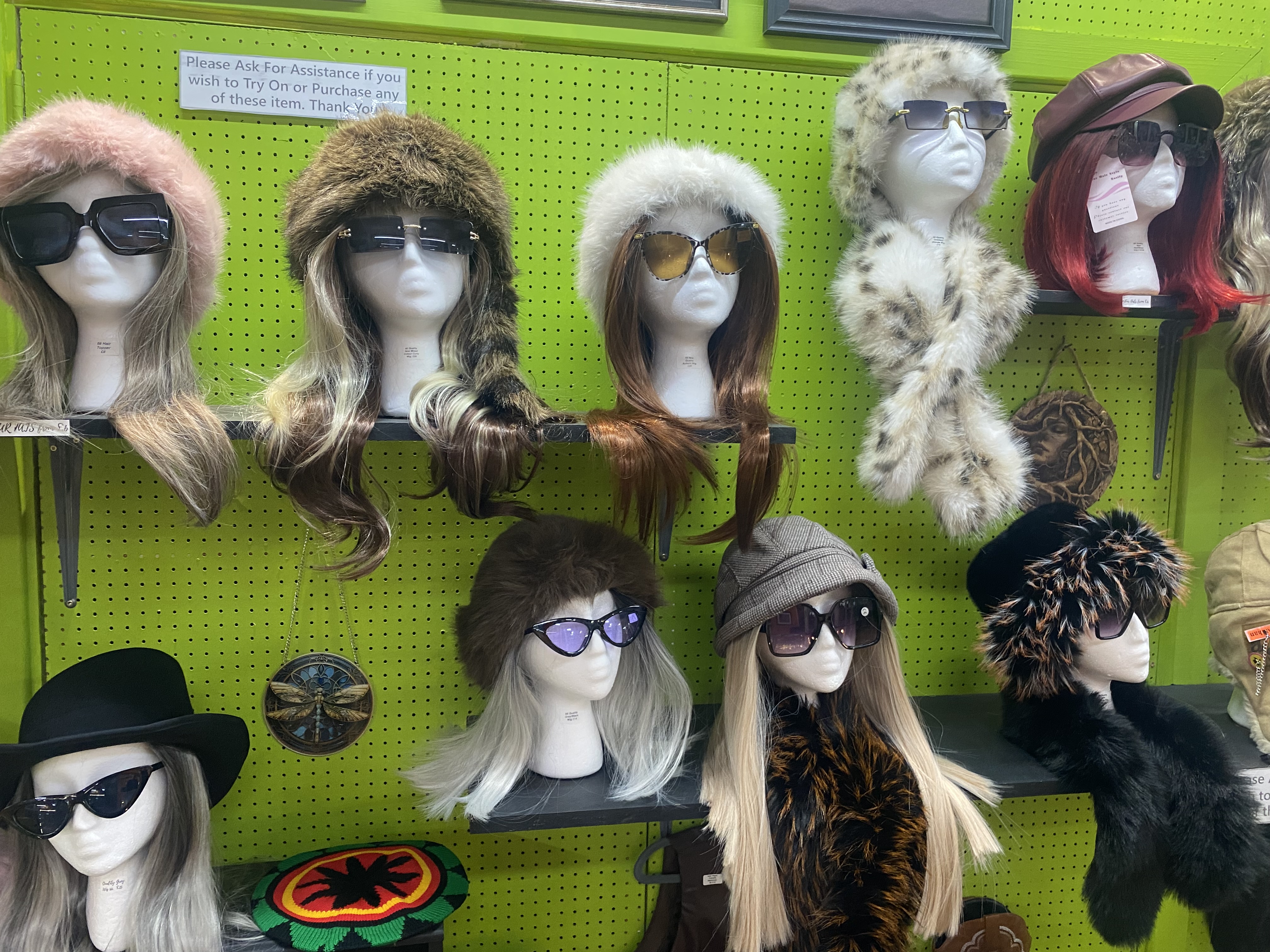 Display of vintage mannequins wearing retro hats, faux-fur accessories, sunglasses, and wigs inside a second-hand shop on bright green pegboard walls.