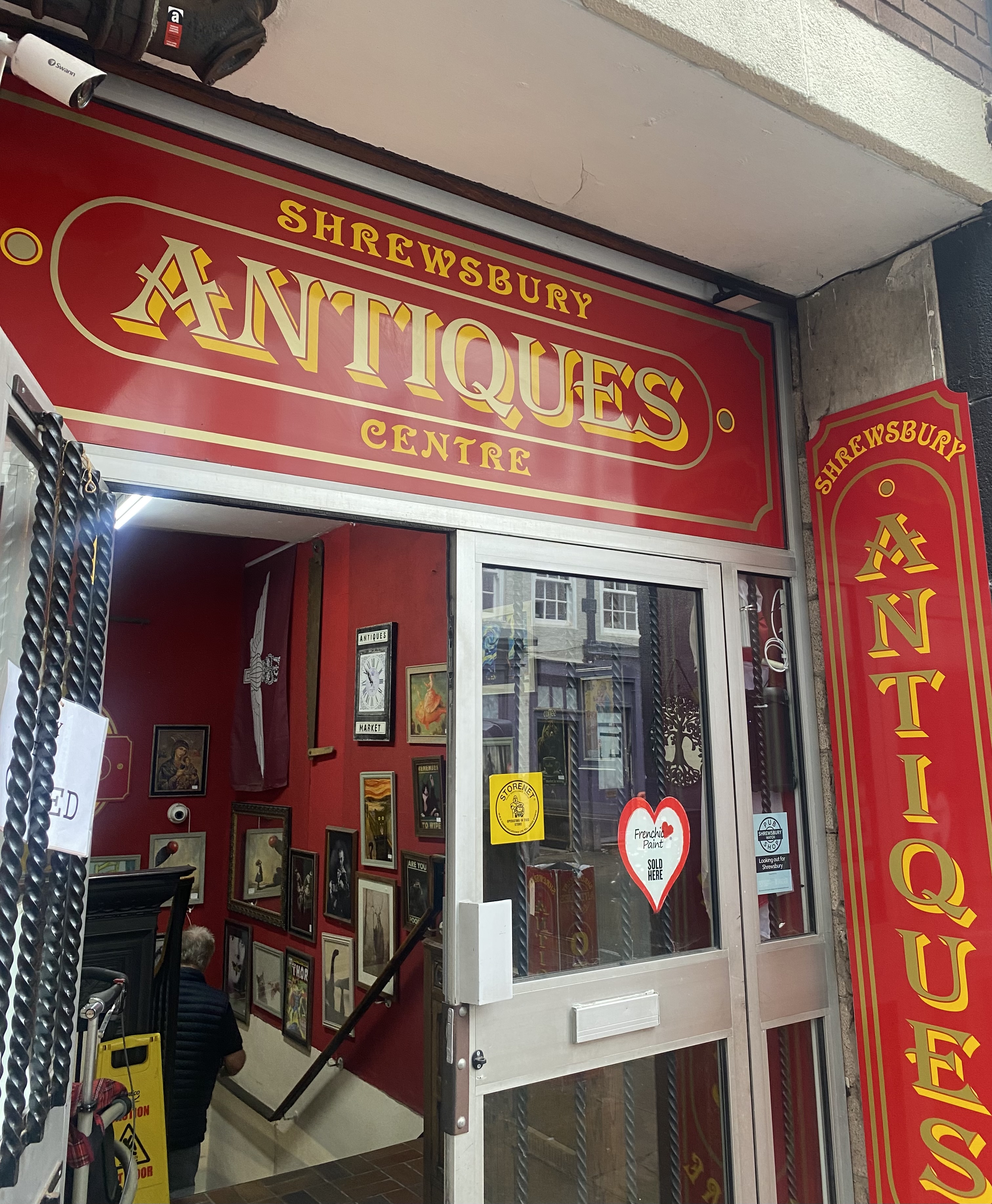 Entrance of Shrewsbury Antiques Centre with red and gold signage, vintage framed artwork displayed inside, and shop window stickers visible.