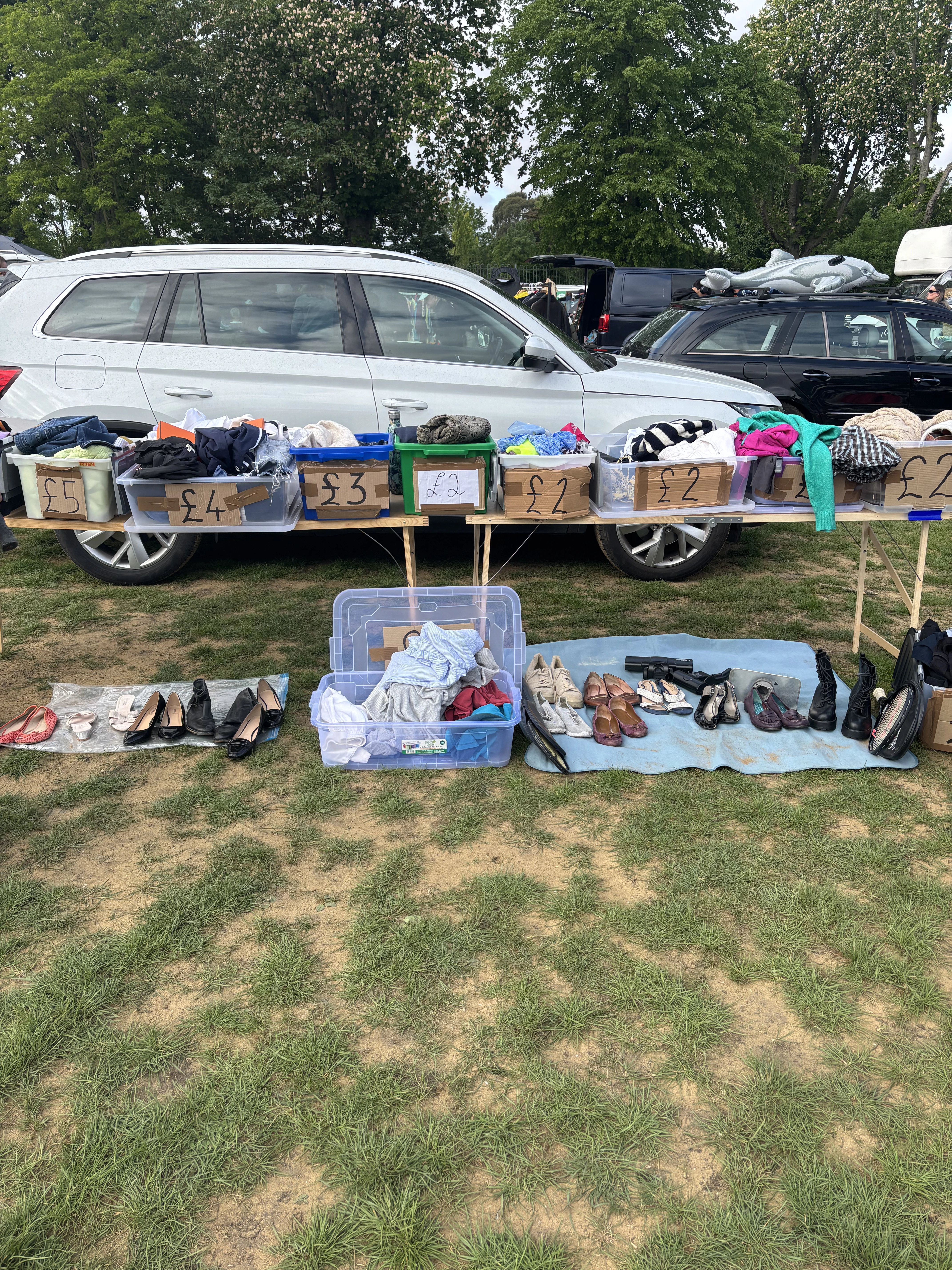 A car boot sale stall on grass in front of parked cars, with a trestle table of clothes sorted into plastic tubs and boxes labelled £5, £4, £3 and £2, and second-hand shoes laid out on blankets on the ground.