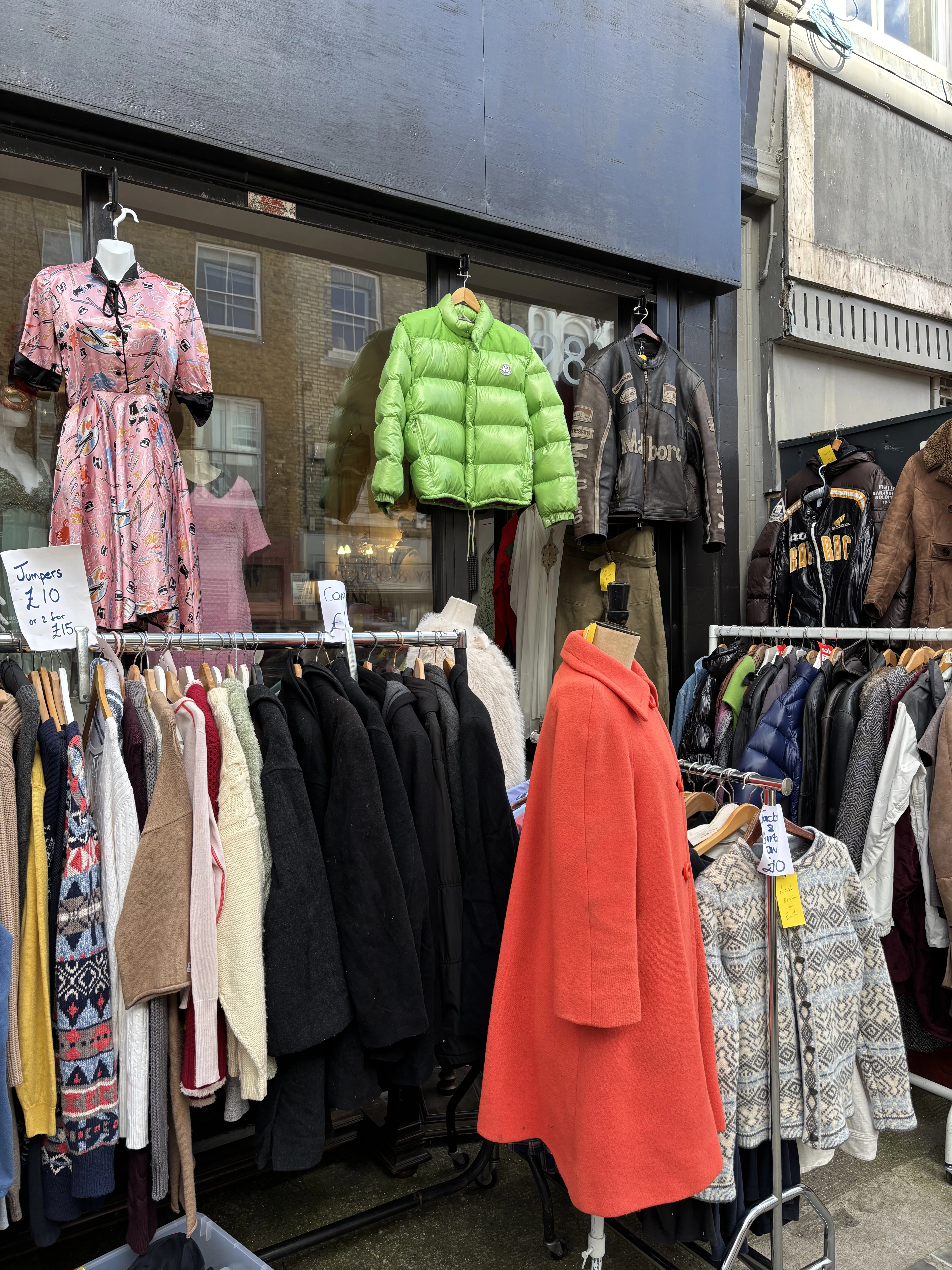 The front of a charity shop with garments hung outside: a patterned pink dress on a mannequin, a lime-green puffer jacket, leather jackets, and rails of coats and knitwear with handwritten price signs reading 'Jumpers £10'.