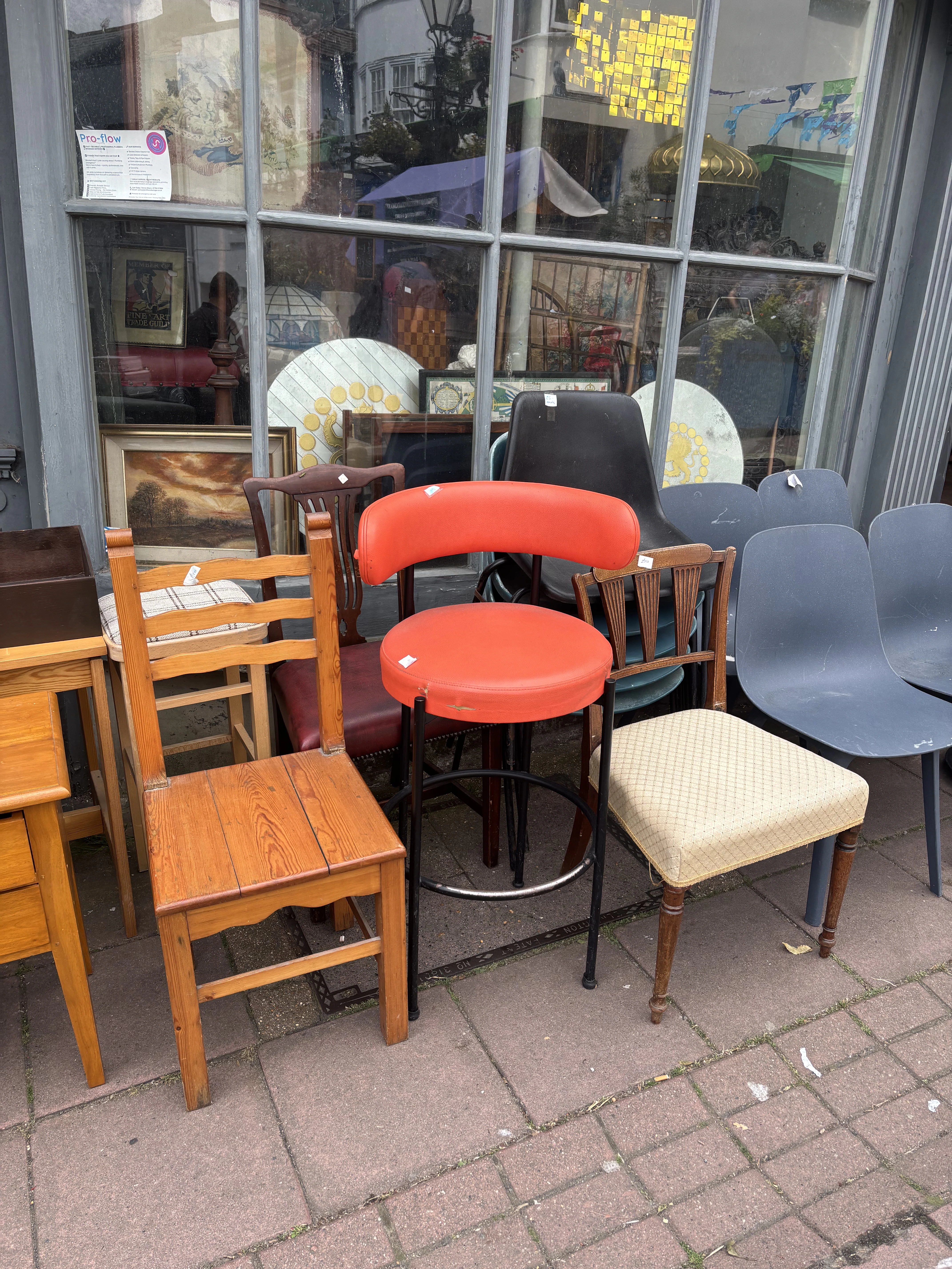 An assortment of second-hand chairs displayed on the pavement outside an indoor flea market, including a pine dining chair, a red vinyl-topped stool, stacked grey plastic chairs and an upholstered chair, with framed paintings and glassware visible in the shop window behind.