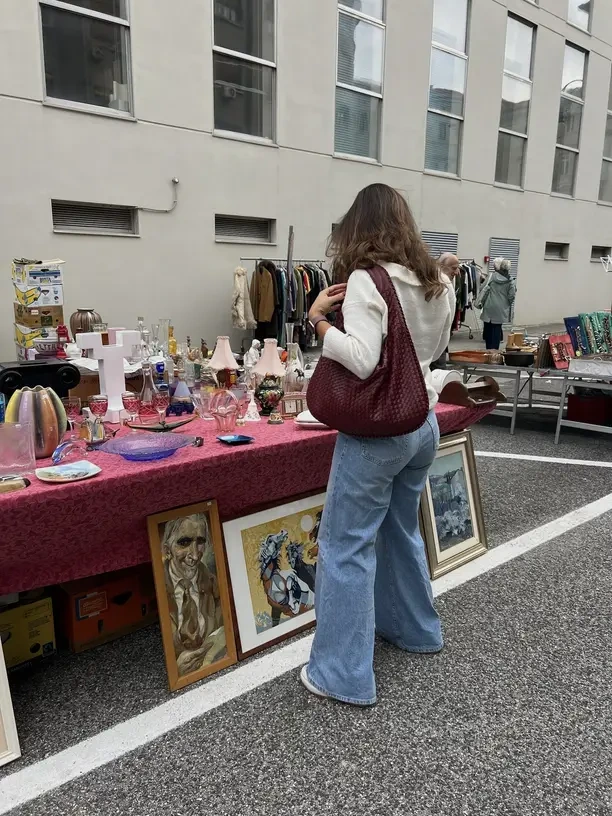 A shopper browses a flea market stall draped with a pink cloth, laid out with vintage glassware, lamps and ceramics, with framed paintings propped against the table and rails of second-hand clothes behind.