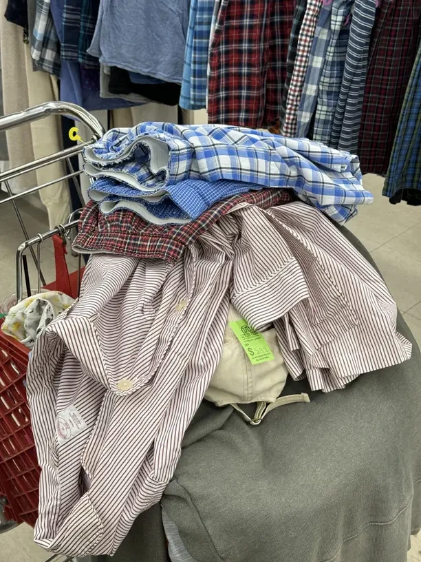 A pile of folded second-hand shirts - blue and red checked patterns and a red pinstripe - stacked on top of a shopping trolley in front of rails of more shirts in a second-hand shop.