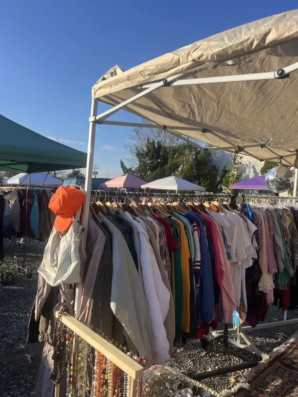 A rail of vintage clothing under a white gazebo at an outdoor flea market, with an orange cap hanging on the end of the rail and rows of colourful garments, jewellery and more market stalls under a clear blue sky.