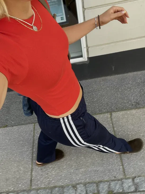 A close-up of an outfit worn on a street: a red ribbed T-shirt with gold necklaces, paired with navy three-stripe track trousers and brown shoes.