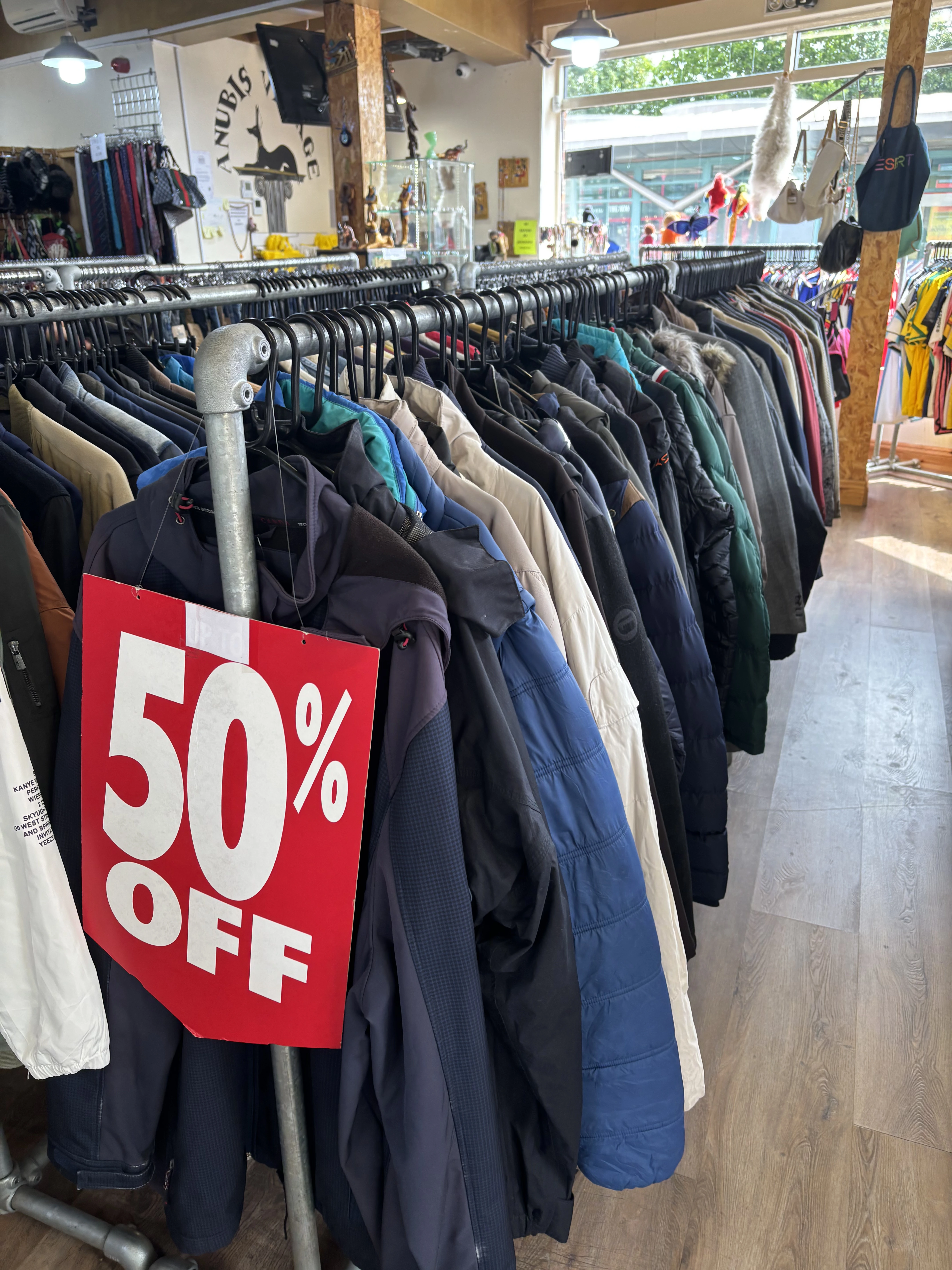 Inside a vintage clothing shop, a long rail packed with coats and jackets in navy, cream, green and black, with a large red '50% OFF' sign hanging at the front and more rails and displays in the background.