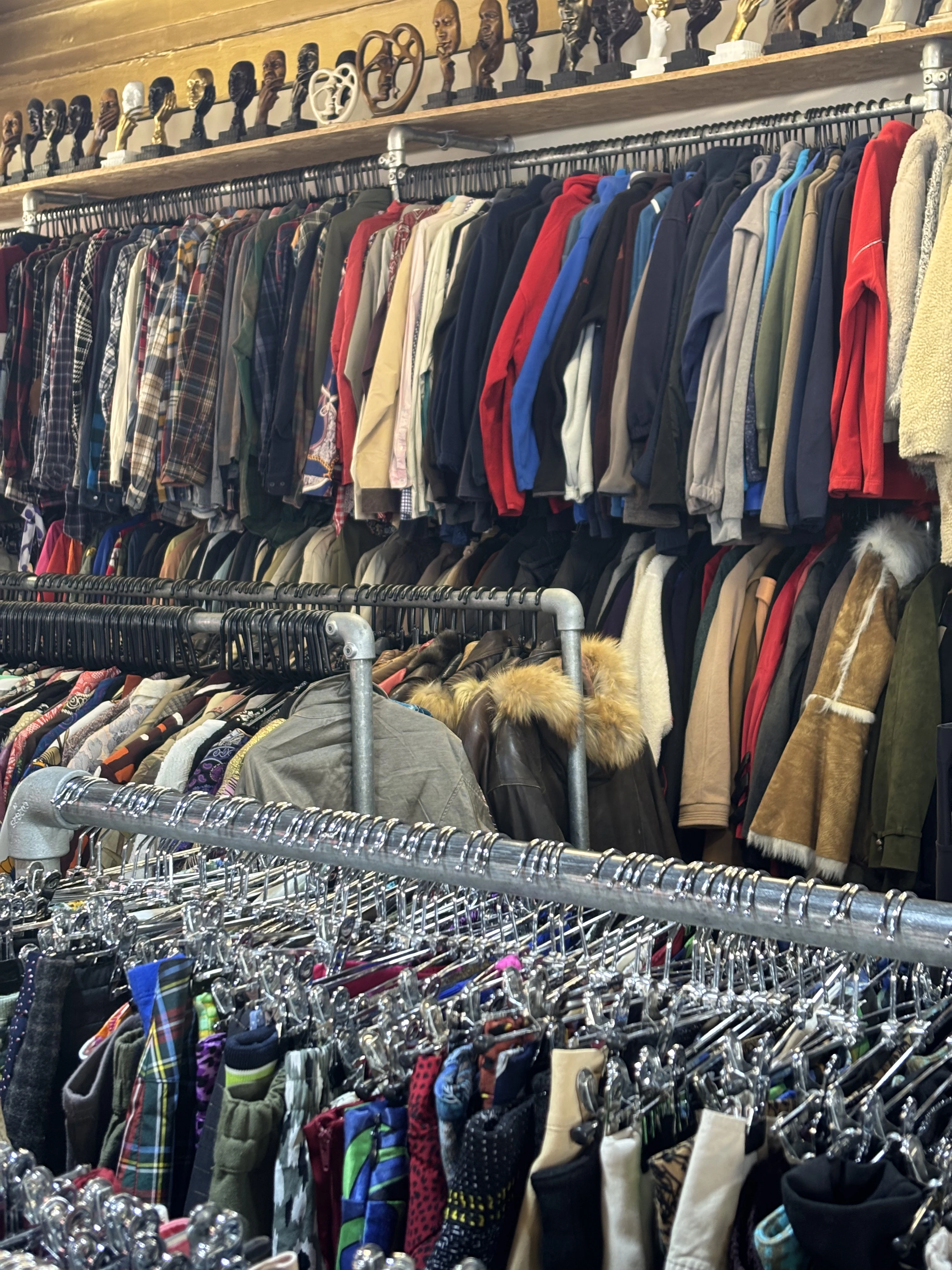The interior of a second-hand clothing shop with densely packed rails of coats, jackets, shirts and fleeces on multiple levels, and a row of carved wooden figurine ornaments displayed on a shelf above.